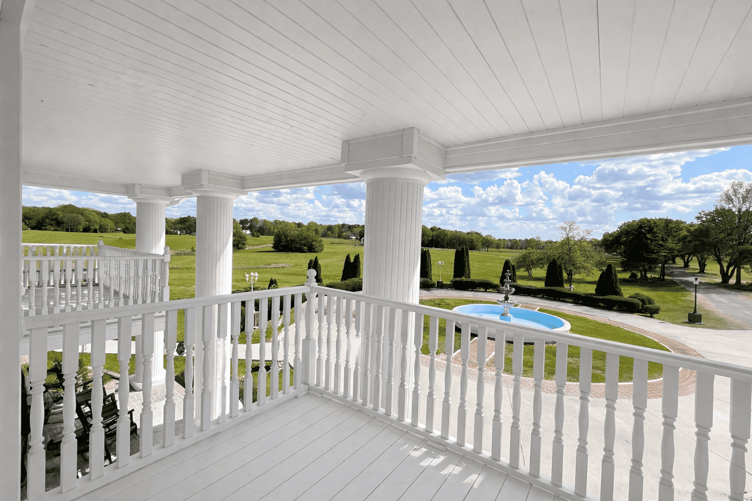 Private balcony at Laurel Haven Estate’s Dogwood Suite featuring white columns and railing, overlooking the fountain, manicured grounds, and expansive countryside views under a bright blue sky.