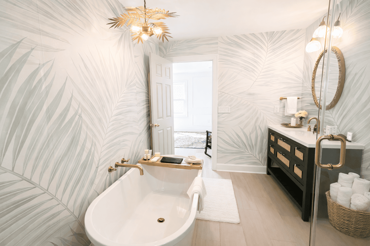 Bright, spa-like bathroom in Laurel Haven Estate’s Dogwood Suite featuring a freestanding soaking tub with bath tray, tropical leaf wallpaper, glass walk-in shower, and styled vanity with fresh flowers, candles, and neatly folded towels.