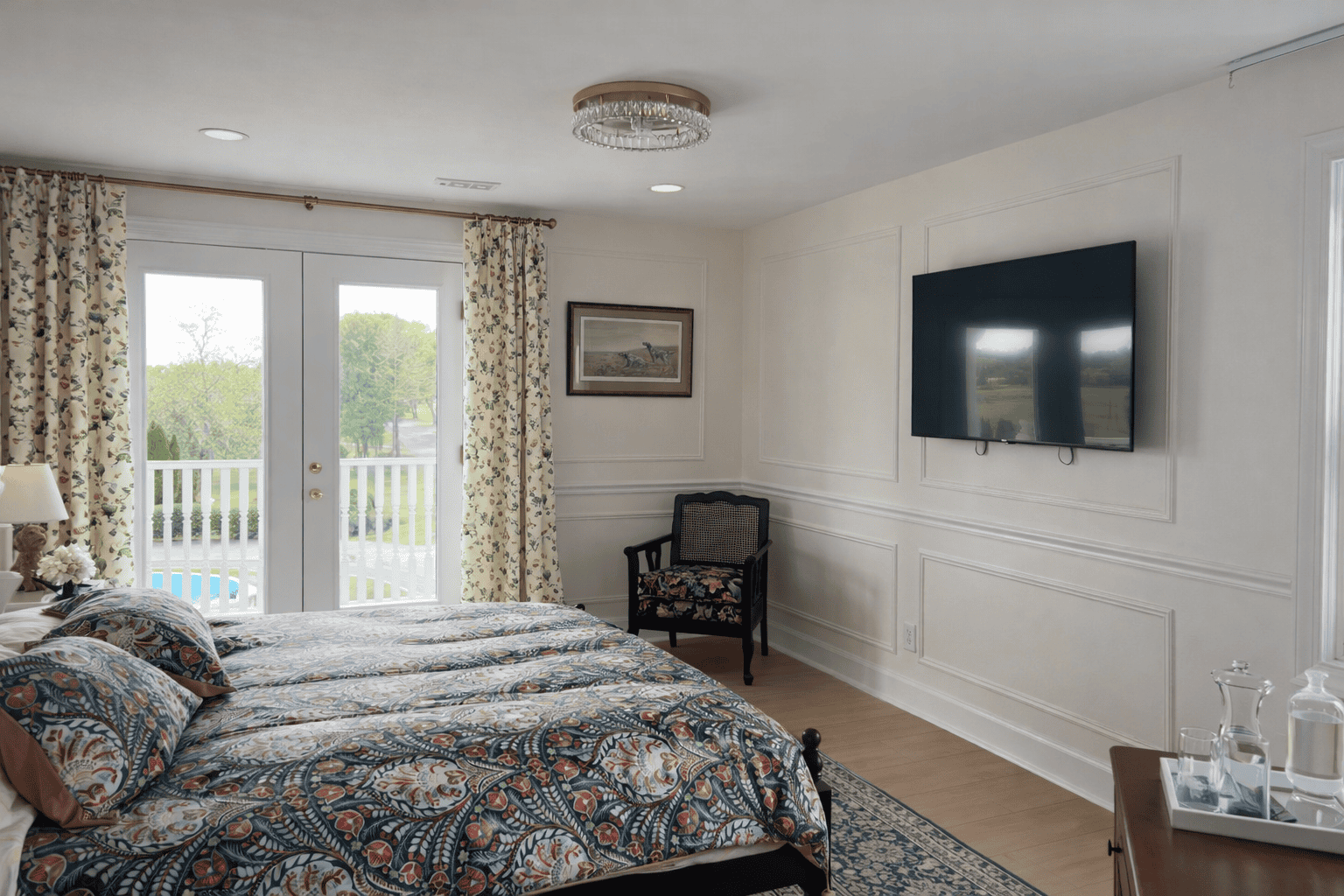 Spacious bedroom in Laurel Haven Estate’s Dogwood Suite featuring a king bed with patterned bedding, wall-mounted TV, elegant trim detail, and French doors with soft patterned curtains opening to a private balcony overlooking the grounds.