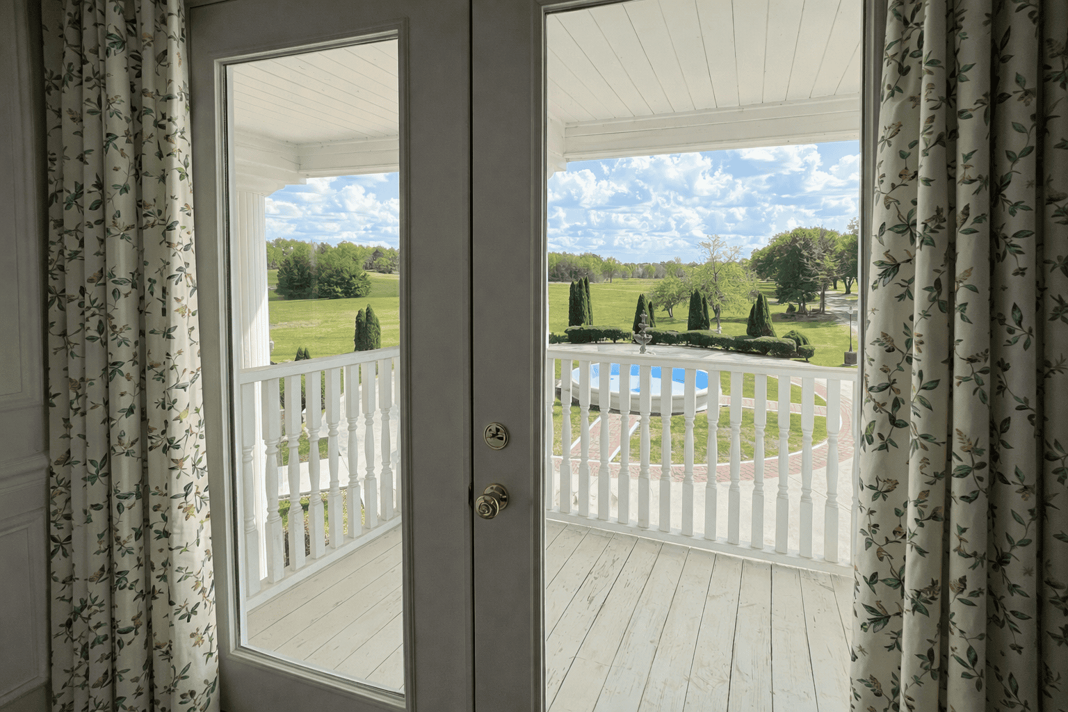 View from inside Laurel Haven Estate’s Dogwood Suite through French doors with soft patterned curtains, opening to a private balcony overlooking the pool, manicured grounds, and scenic countryside.