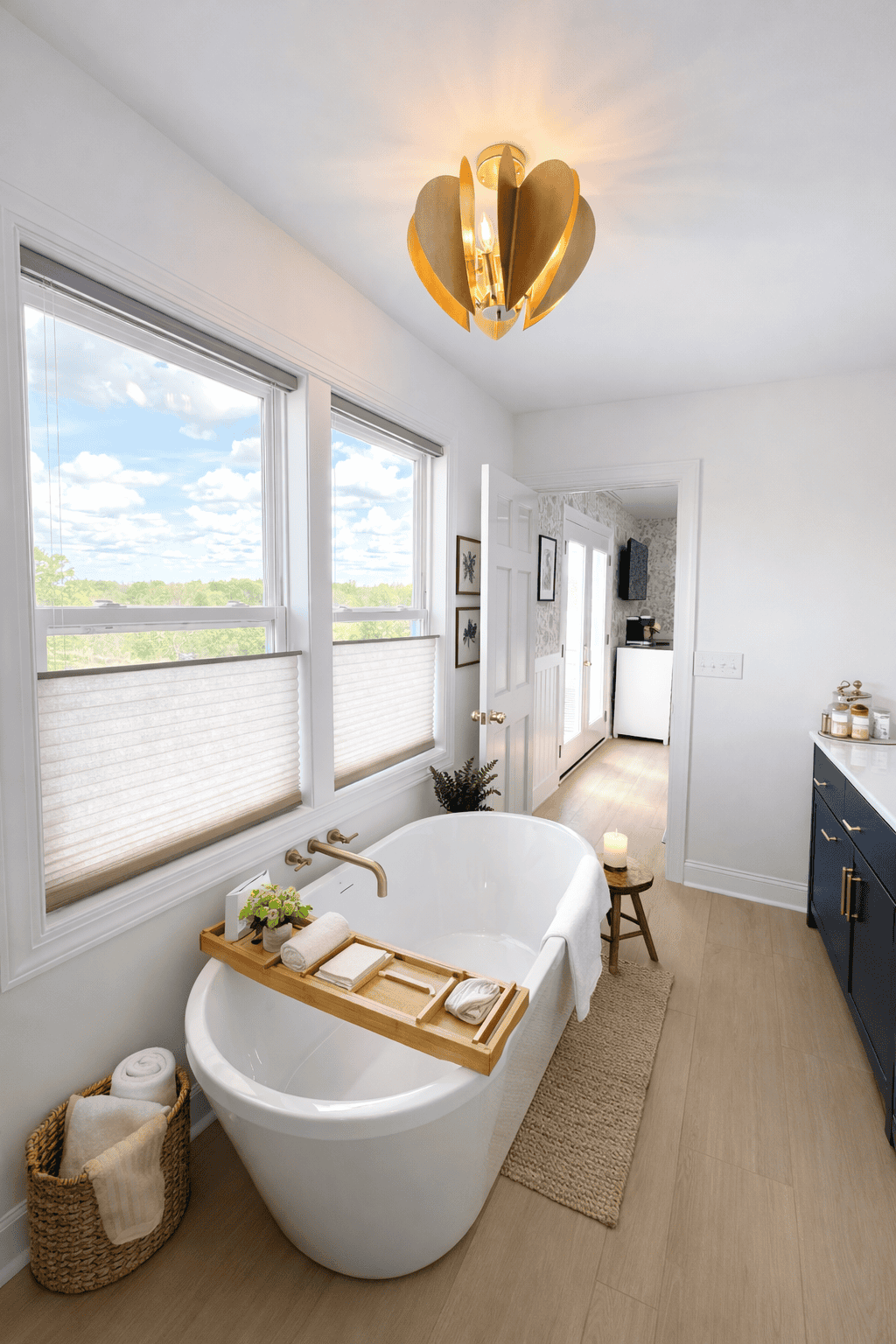 Bright, spa-style bathroom with a freestanding soaking tub beneath large windows overlooking scenic vineyard views, styled with a wooden bath tray, rolled towels, greenery, and neutral decor, featuring a navy vanity and warm wood flooring.