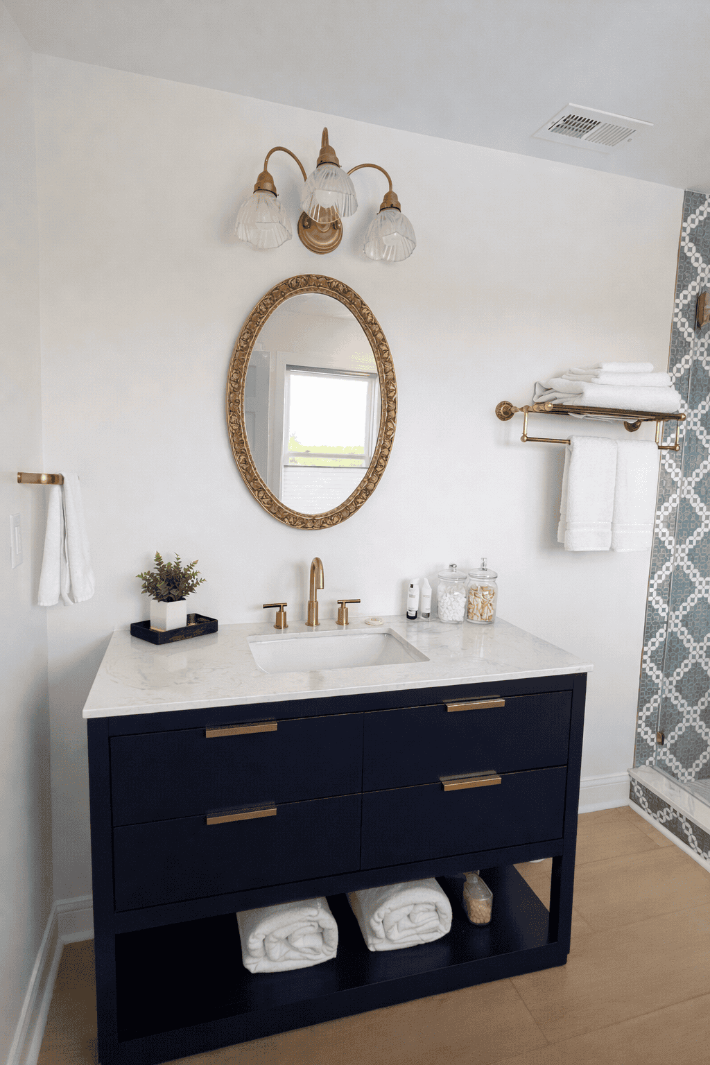 Bright, elegant bathroom in the Jessamine Suite featuring a navy-blue vanity with a white marble countertop, gold fixtures, and an ornate oval mirror, styled with fresh greenery, rolled towels, and glass apothecary jars, alongside a geometric tile walk-in shower for a clean, upscale, real estate–ready presentation.