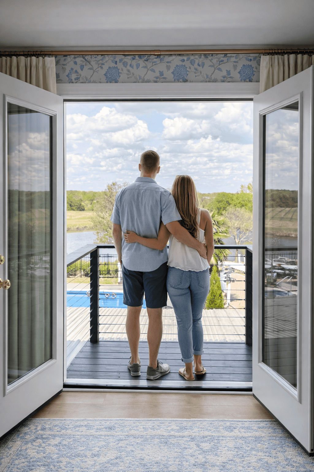 Couple standing on Romeo and Juliet balcony at Laurel Haven Estate in Lancaster SC overlooking vineyard, pond, and pool through open French doors with soft curtains