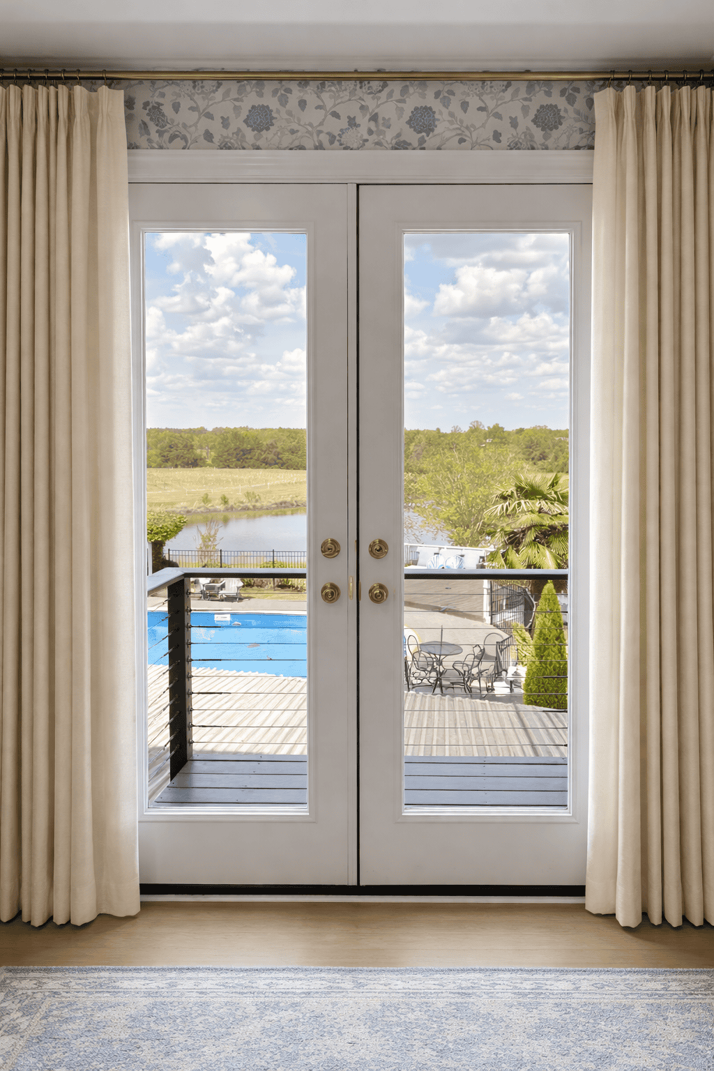 View through French doors in Jessamine Suite at Laurel Haven Estate in Lancaster SC showing balcony, pool, pond, and vineyard with soft curtains framing the scene