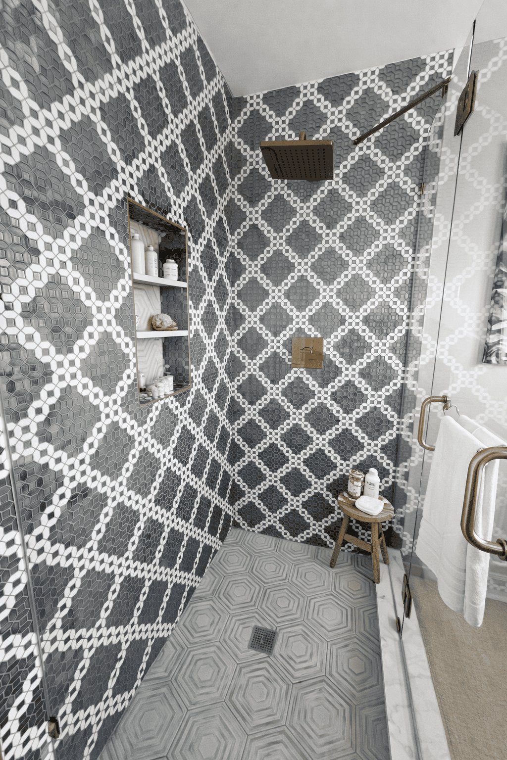 Bright, modern walk-in shower with glass enclosure, bold gray and white geometric tile walls, brass rainfall showerhead and fixtures, styled with spa accessories, folded towels, and a small wooden stool for a clean, real estate–ready look.